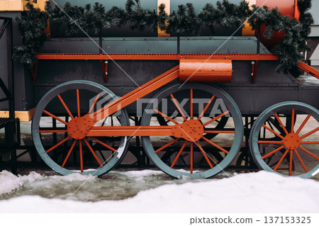Close-up of red wheels of a decorative vintage steam train decorated with pine garlands in winter. Industrial holiday background with snow. Close-up of red wheels of a decorative vintage steam train decorated with pine garlands in winter. Industrial holiday background with snow. 137153325