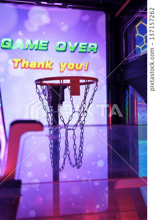 Close-up of arcade basketball hoop with chain net and illuminated Game Over screen in the background. Neon entertainment atmosphere in gaming center. 137157262