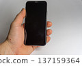 Hand holds a smartphone with a dark screen in a plain white setting during daytime 137159364