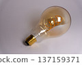 Light bulb with visible filament sits on a plain surface in a well lit area during daylight 137159371