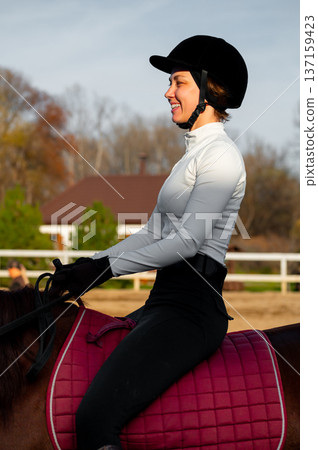 Female equestrian in black riding attire and helmet sits on a brown horse with a pink saddle pad, outdoor riding arena and trees visible in the background 137159423