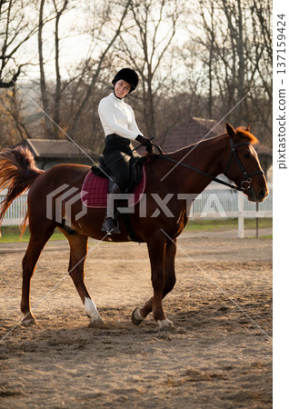 horse rider looks at the camera and smiles in an open arena nearby, sitting in the saddle and wearing a helmet. Horse training and skill development horse rider looks at the camera and smiles in an open arena nearby, sitting in the saddle and wearing a helmet. Horse training and skill development 137159424