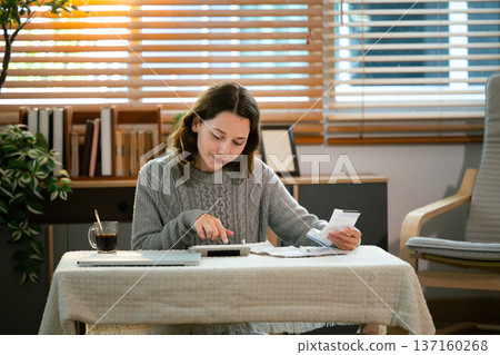 Caucasian woman using calculator managing household expenses and taxes in a living room 137160268