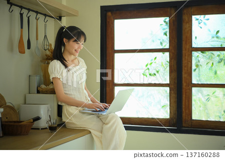 Young woman uses laptop sitting on kitchen counter Young woman uses laptop sitting on kitchen counter 137160288