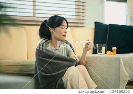 Young woman wrapped in blanket checks temperature while resting indoors Young woman wrapped in blanket checks temperature while resting indoors 137160293