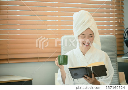 Young woman in bathrobe reads book while drinking tea indoors Young woman in bathrobe reads book while drinking tea indoors 137160324
