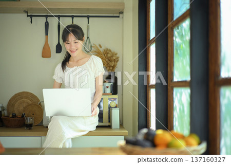 Young woman uses laptop sitting on kitchen counter 137160327