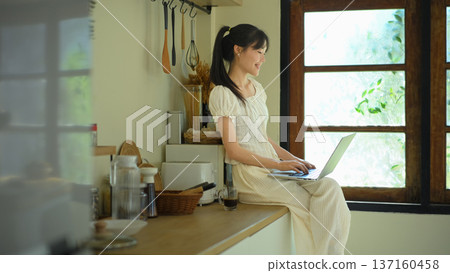 Young woman uses laptop sitting on kitchen counter Young woman uses laptop sitting on kitchen counter 137160458