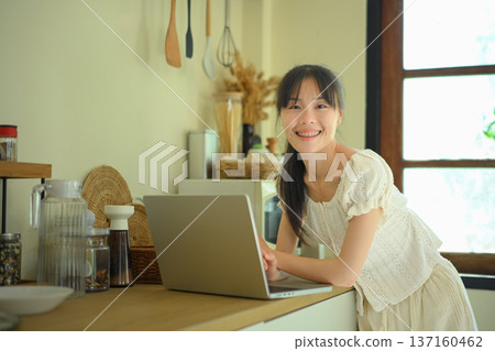 Young woman while working on laptop in kitchen and looking at camera in kitchen 137160462