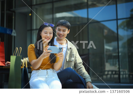 Asian couple relaxing outdoors after shopping, smiling while checking a smartphone together beside colorful shopping bags Asian couple relaxing outdoors after shopping, smiling while checking a smartphone together beside colorful shopping bags 137160506
