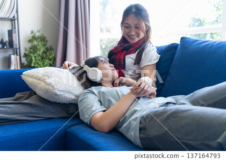 Cozy Relaxation Moment: Two individuals share an intimate moment on a comfortable sofa. One lies relaxed, listening to music through headphones, while the other watches over them. Cozy Relaxation Moment: Two individuals share an intimate moment on a comfortable sofa. One lies relaxed, listening to music through headphones, while the other watches over them. 137164793