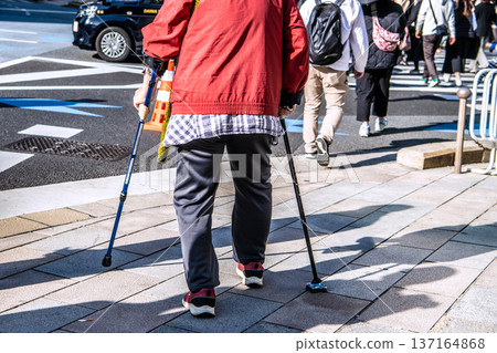 Tokyo cityscape, Japan. An elderly woman with a walking stick.... On the sidewalk in front of Mitsukoshimae Station in Nihonbashi on the day of the Tokyo Marathon. Tokyo cityscape, Japan. An elderly woman with a walking stick.... On the sidewalk in front of Mitsukoshimae Station in Nihonbashi on the day of the Tokyo Marathon. 137164868