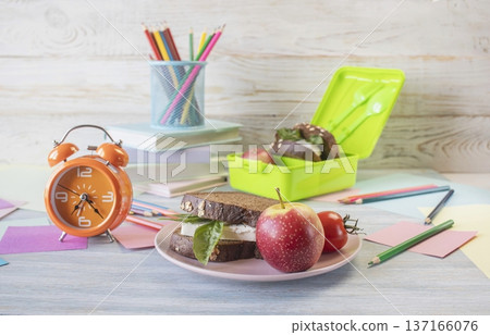 Back to school concept, school food. Lunch box with healthy sandwich, alarm clock, stack of books and apple on wooden background. Back to school concept, school food. Lunch box with healthy sandwich, alarm clock, stack of books and apple on wooden background. 137166076