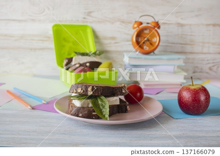 Back to school concept, school food. Lunch box with healthy sandwich, alarm clock, stack of books and apple on wooden background. Back to school concept, school food. Lunch box with healthy sandwich, alarm clock, stack of books and apple on wooden background. 137166079