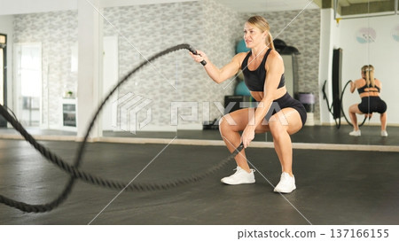 Woman exercising with battle ropes in gym squatting for fitness 137166155