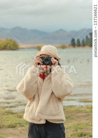 Woman in cozy jacket capturing the moment in autumn nature with retro camera by the lake at New Zealand 137166621