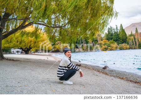 Young asian woman crouching under willow tree by lakeshore and flock of duck in autumn Young asian woman crouching under willow tree by lakeshore and flock of duck in autumn 137166636