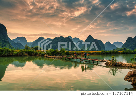 Sunset over Phong Nam Valley with rice fields and farmers crossing wooden bridge in countryside 137167114