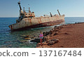 Woman standing on the rocky seashore admiring the iconic rust-covered Edro III Shipwreck grounded on the coast of Paphos, Cyprus, with clear blue Mediterranean sea and sky 137167484