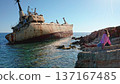 Young woman sitting on a rocky seashore admiring the iconic Edro III shipwreck, a popular tourist landmark near Paphos, under a bright blue sky on a clear day 137167485