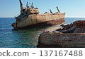 Young Caucasian woman sitting on coastal rocks, looking at the rusty Edro III shipwreck partially submerged in the clear turquoise waters of the Mediterranean Sea near Paphos, Cyprus 137167488