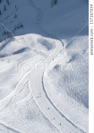 Winding ski slope on snowy mountain in Krasnaya Polyana shows groomed track with skiers descending white alpine terrain under clear winter sky, Winter tourism and outdoor sport concept for travel Winding ski slope on snowy mountain in Krasnaya Polyana shows groomed track with skiers descending white alpine terrain under clear winter sky, Winter tourism and outdoor sport concept for travel 137167834