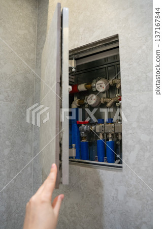 Hand opening wall cabinet with water meters and pipe system reveals residential plumbing infrastructure with gauges and valves, Home maintenance, utility management and resource monitoring Hand opening wall cabinet with water meters and pipe system reveals residential plumbing infrastructure with gauges and valves, Home maintenance, utility management and resource monitoring 137167844