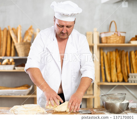 Male baker in unbuttoned uniform prepares dough, kneads it with his hands Male baker in unbuttoned uniform prepares dough, kneads it with his hands 137170801