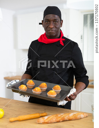 Proffesional man in black chef uniform holding sheet pan with just baked cupcakes Proffesional man in black chef uniform holding sheet pan with just baked cupcakes 137170802