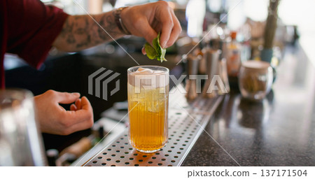 Serving drink. Barkeeper work. Tattooed hand adding green mint leaves to yellow refreshing cocktail in glass with ice cubes whipped cream. Serving drink. Barkeeper work. Tattooed hand adding green mint leaves to yellow refreshing cocktail in glass with ice cubes whipped cream. 137171504