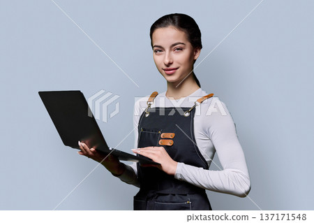 Portrait of young smiling woman in apron holding laptop on gray background Portrait of young smiling woman in apron holding laptop on gray background 137171548