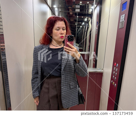 Woman in checked blazer and short brown skirt taking a selfie in elevator close-up. Expression, individuality, and urban confidence captured in a casual moment of everyday life. Woman in checked blazer and short brown skirt taking a selfie in elevator close-up. Expression, individuality, and urban confidence captured in a casual moment of everyday life. 137173459
