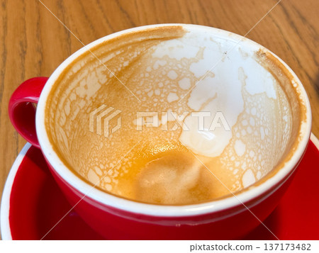 Empty coffee cup with foam traces on red saucer close-up. Warmth, everyday ritual and comfort after morning drink in cozy cafe. 137173482