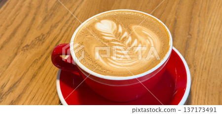 Cup of cappuccino with latte art in red cup on wooden table banner. Warmth, aroma, and comfort in cozy cafe atmosphere. Cup of cappuccino with latte art in red cup on wooden table banner. Warmth, aroma, and comfort in cozy cafe atmosphere. 137173491