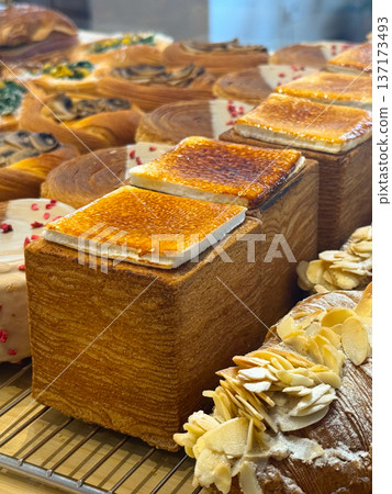 Close-up Modern bakery display with cube pastries topped with caramelized sugar and round buns with white icing and dried berries. Confectionery precision and artistic presentation. Close-up Modern bakery display with cube pastries topped with caramelized sugar and round buns with white icing and dried berries. Confectionery precision and artistic presentation. 137173493