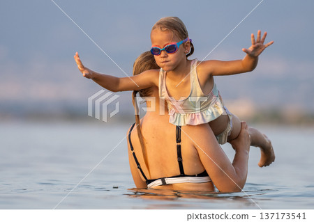 Woman lifting little girl in swimming goggles above sea water during summer vacation at the beach 137173541
