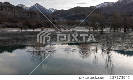 Wide angle view of a reservoir with frozen edges and snow covered peaks under a cloudy sky Wide angle view of a reservoir with frozen edges and snow covered peaks under a cloudy sky 137173945