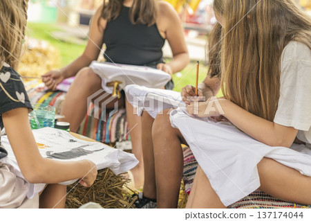 Young girls draw on white t-shirts with pencils and paint, outdoor art workshop sitting together on hay bales in summer park. Creativity, DIY fashion, children activity, handmade 137174044