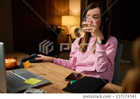 Woman wearing headphones drinks wine while sitting at desk with laptop in home interior Woman wearing headphones drinks wine while sitting at desk with laptop in home interior 137174341