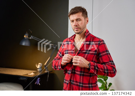 Man standing indoors and buttoning plaid shirt Man standing indoors and buttoning plaid shirt 137174342