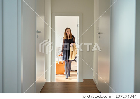 Woman in dress holding coat and suitcase walking through hallway 137174344