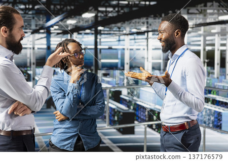 Team of software engineers working on AI automatization in data center, standing on industrial platform. Server farm employees collaborating for server management with artificial intelligence tools. Team of software engineers working on AI automatization in data center, standing on industrial platform. Server farm employees collaborating for server management with artificial intelligence tools. 137176579