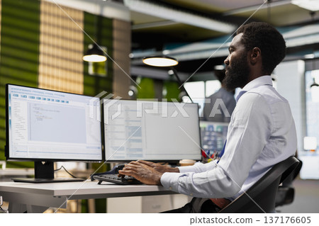 Software developer reviewing code snippets in modern workplace, adjusting configurations. African american man at work running diagnostic tools on PC, monitoring API cloud services 137176605
