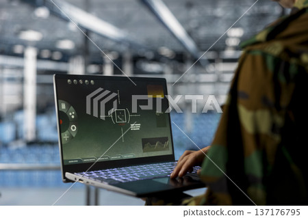 Close up of army officer in camouflage uniform operating laptop with missile lock interface inside modern data center. Military professional tracking targets using notebook dashboard 137176795