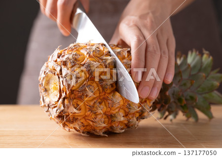 Close up of hand holding kitchen knife and cutting pineapple on wooden board Close up of hand holding kitchen knife and cutting pineapple on wooden board 137177050