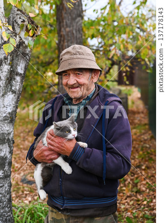 Elderly Russian man standing outdoors with his gray and white cat in his arms on an autumn day, smiling kindly and enjoying peaceful rural life. 137178143