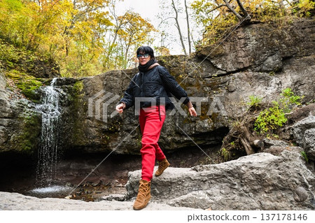 Active 50 year old Russian woman hiking near a small waterfall in the mountains of Primorsky Krai, Russia. She walks on rocks with a backpack outdoors. Active 50 year old Russian woman hiking near a small waterfall in the mountains of Primorsky Krai, Russia. She walks on rocks with a backpack outdoors. 137178146