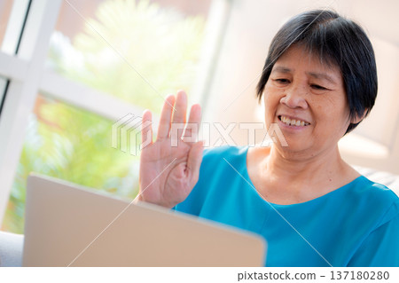 Senior asian woman waving during video call on laptop computer sitting on sofa in living room. Senior asian woman waving during video call on laptop computer sitting on sofa in living room. 137180280