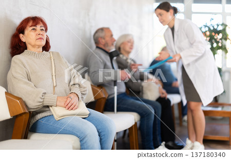 Group of elderly people sitting in chairs in queue to see doctor, nurse talking with patients in background in lobby medical facility Group of elderly people sitting in chairs in queue to see doctor, nurse talking with patients in background in lobby medical facility 137180340