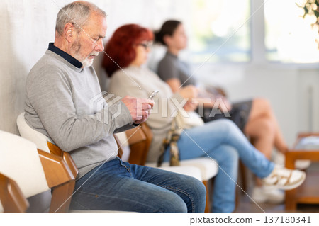 Elderly man sitting in clinic waiting area scrolling mobile phone Elderly man sitting in clinic waiting area scrolling mobile phone 137180341